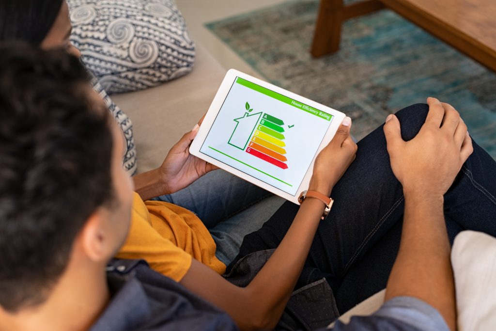 O persoană care ține în mână o tabletă care arată o diagramă de evaluare a eficienței energetice cu o pictogramă a casei și niveluri cu coduri de culori, de la roșu la verde, stă pe o canapea bej. Alături de ei, un alt individ se angajează într-un duel pro și contra despre beneficiile vată minerală versus Spuma poliuretanică, cu o parte dintr-o măsuță de cafea din lemn vizibilă.