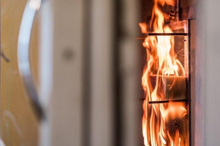 Prim-plan al unei sobe pe lemne cu o ușă de sticlă, care arată flăcările care ard activ în interior. Imaginea se concentrează asupra focului și a unei părți din structura sobei, subliniind mânerul din partea stângă. Proiectat având în vedere siguranța, evidențiază măsuri împotriva inflamabilității pentru a asigura o utilizare în siguranță.