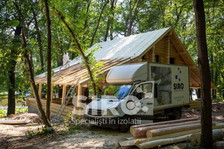 Casă din lemn în lucru, cu autoutilitara echipei lângă șantier